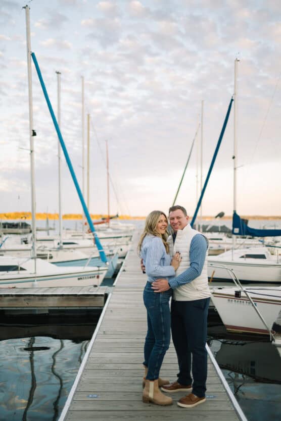 A couple stands close together, smiling, on a dock lined with sailboats at a marina during sunset. The sky is partly cloudy and the water is calm, creating a peaceful and romantic atmosphere. by Justin Salem Meyer