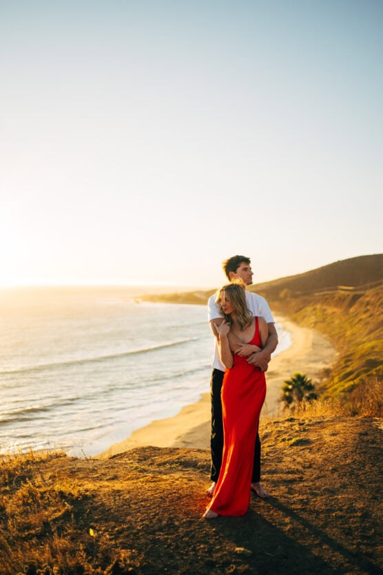 A couple stands on a cliff overlooking the ocean at sunset. The woman in a red dress hugs the man from behind as they gaze toward the horizon, with a sandy beach and hills in the background, bathed in golden light. by Justin Salem Meyer