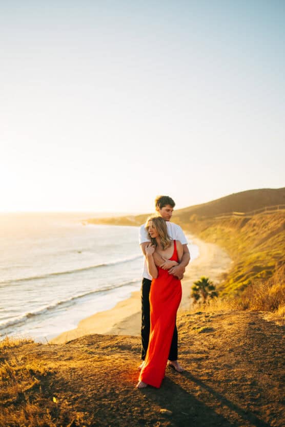 A couple stands on a cliff overlooking the ocean at sunset. The woman wears a bright red dress while the man, in a white shirt, embraces her from behind with rolling hills and a sandy beach in the background. by Justin Salem Meyer
