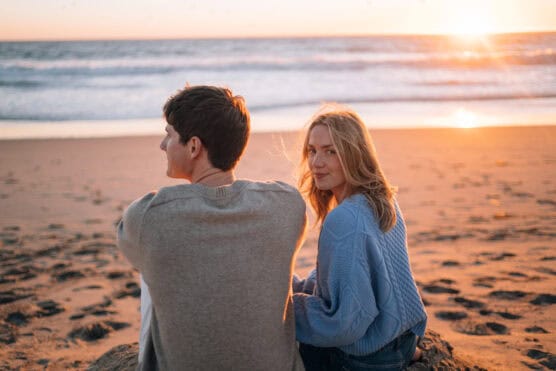 A young couple sits on the sand at the beach during sunset. The man faces the ocean while the woman, in a blue sweater, turns to look toward the camera. The sun sets over the water in the background. by Justin Salem Meyer
