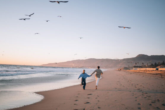 A couple holds hands while running along a sandy beach at sunset, with seagulls flying overhead and gentle waves lapping the shore; mountains and coastal buildings are visible in the background. by Justin Salem Meyer