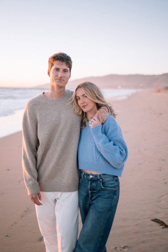 A couple stands on a sandy beach at sunset. The man wears a beige sweater and white pants, and the woman wears a light blue sweater and jeans, with her arm around his shoulders. The ocean and distant hills are in the background. by Justin Salem Meyer
