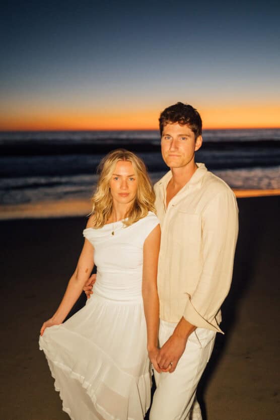 A couple stands on a beach at sunset, holding hands. The woman wears a white dress and the man wears a light shirt and white pants. The sky behind them is deep blue and orange, with waves visible in the background. by Justin Salem Meyer