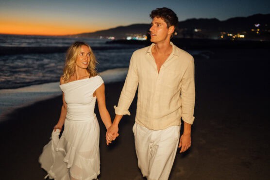 A couple walks hand in hand along a beach at sunset. The woman wears a white dress, and the man wears a light-colored shirt and pants. The sky is orange and purple, and waves are visible in the background. by Justin Salem Meyer