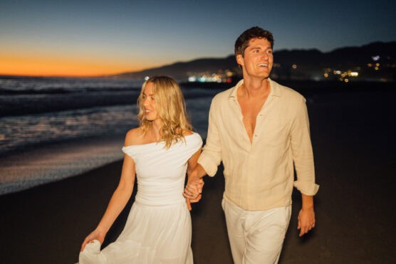 A smiling couple walks hand in hand along a beach at sunset. The woman wears a white off-shoulder dress, and the man wears a light-colored shirt and pants. The sky is a gradient of orange to deep blue. by Justin Salem Meyer