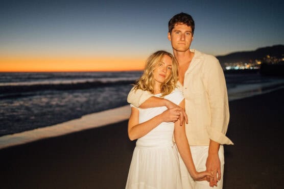 A couple stands close together on a beach at sunset, dressed in white; the sky glows orange and blue, and gentle waves are visible in the background. by Justin Salem Meyer