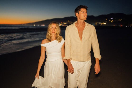 A couple dressed in light, summery clothing walks hand-in-hand along a beach at sunset, with the ocean and distant lights visible behind them. by Justin Salem Meyer