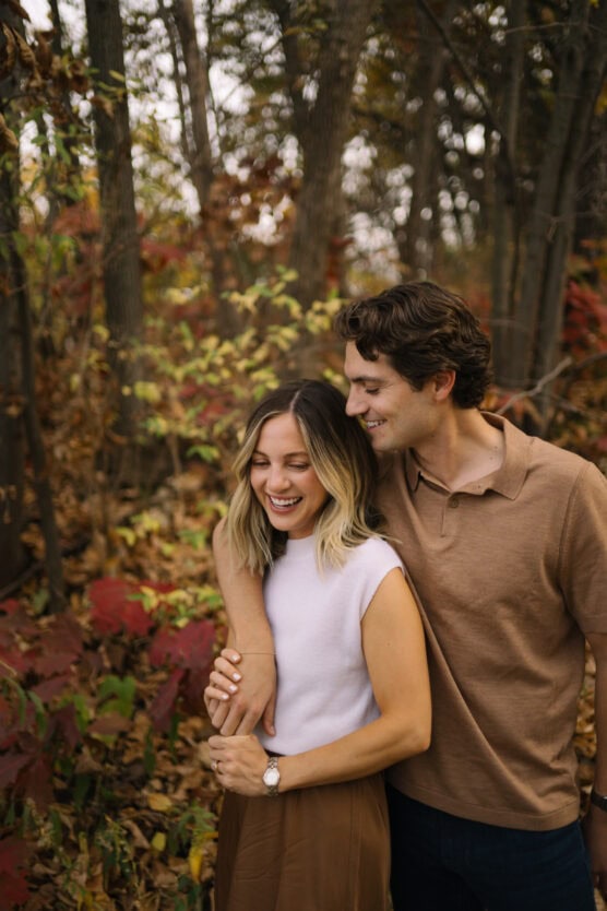 A young couple stands close together in a forest during autumn. The woman is smiling and laughing while the man hugs her from the side. Colorful fall leaves surround them. by Justin Salem Meyer