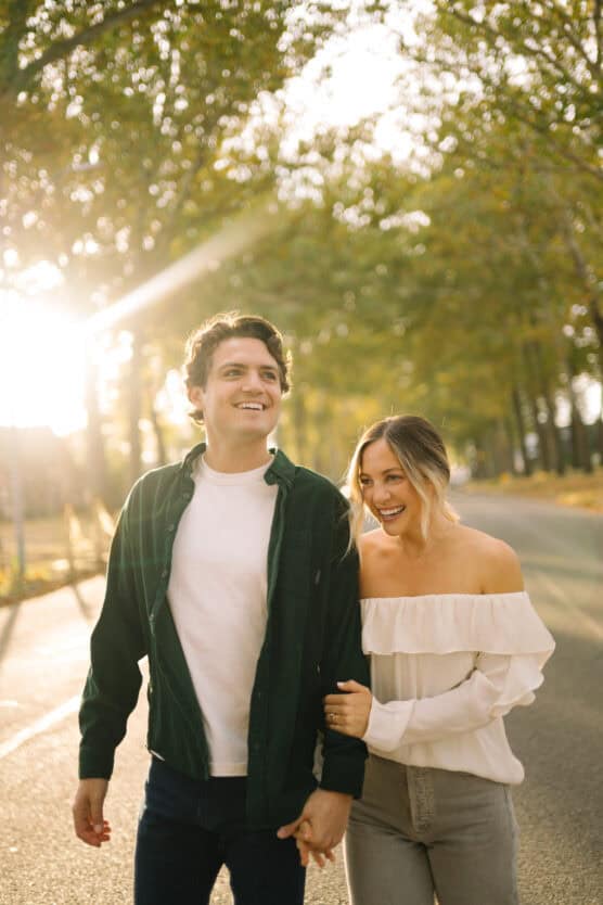 A young couple walks on a sunlit road lined with trees, smiling and holding hands. The woman wears an off-shoulder white top; the man wears a white shirt and dark jacket. Sunlight filters through the trees in the background. by Justin Salem Meyer