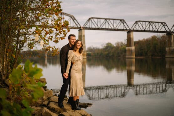 A smiling couple stands on rocky ground by a calm river, holding hands. Behind them, trees with autumn leaves frame the scene, and an old railroad bridge spans the water under a cloudy sky. by Justin Salem Meyer
