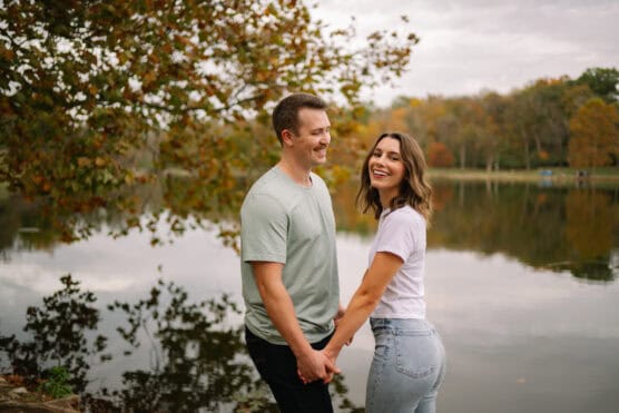 A smiling couple holds hands and faces each other by a calm lake, with autumn trees reflected in the water in the background. The sky is overcast, and both are casually dressed. by Justin Salem Meyer
