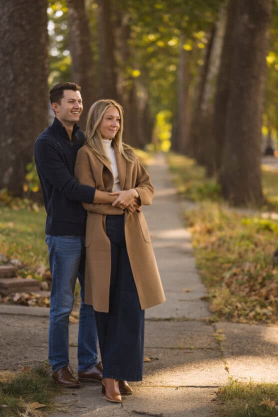 A man and woman stand on a tree-lined sidewalk. The man, wearing a dark sweater and jeans, stands behind the woman, embracing her. She wears a tan coat and dark pants. They both look ahead, smiling, surrounded by autumn foliage. by Justin Salem Meyer