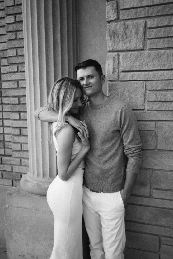 A couple stands close together in front of a stone wall with columns. The man, in a sweater and pants, leans against the wall, smiling slightly. The woman, wearing a light dress, rests her head on his chest and holds his hand. by Justin Salem Meyer