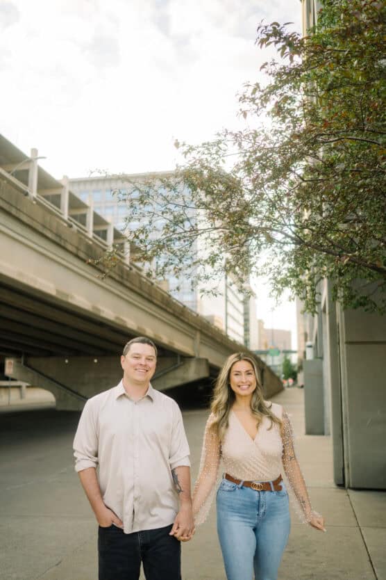 A smiling couple holds hands and stands on a city sidewalk beneath an overpass, with buildings and tree branches visible in the background. by Justin Salem Meyer