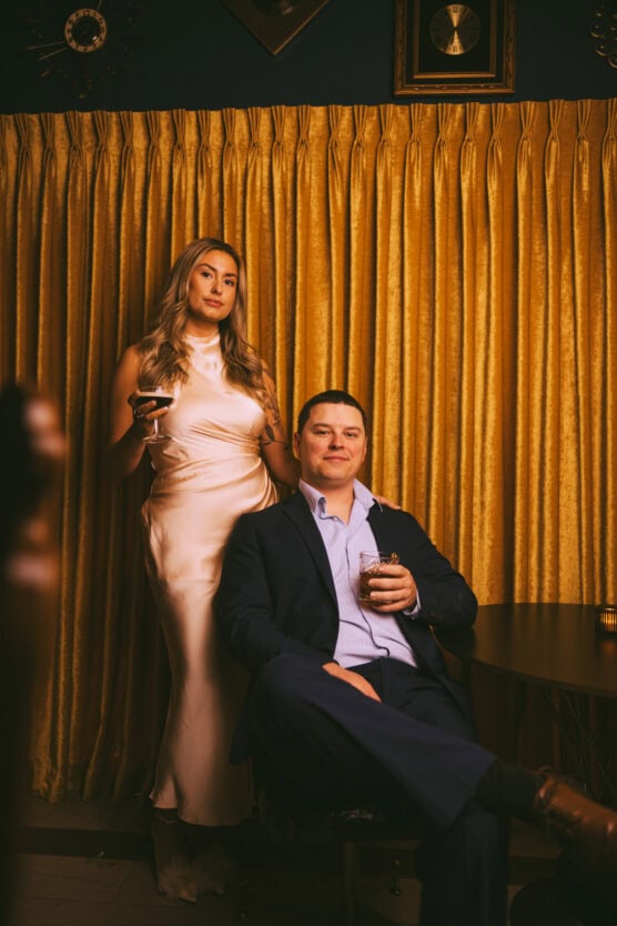 A woman in a silky, light-colored dress stands holding a drink beside a seated man in a dark suit with a drink in hand. They pose in front of gold curtains, with framed decor and a clock on the wall behind them. by Justin Salem Meyer