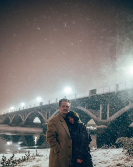 A man and woman stand together in a snowy landscape at night near a river, with the woman resting her head on the mans shoulder. A lit bridge arches in the background amid falling snow. by Justin Salem Meyer