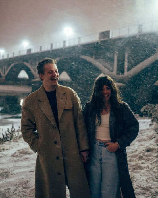 A man and woman stand hand in hand, smiling in the snow at night. They wear long coats and are near a bridge lit by streetlights, with snow gently falling around them. by Justin Salem Meyer
