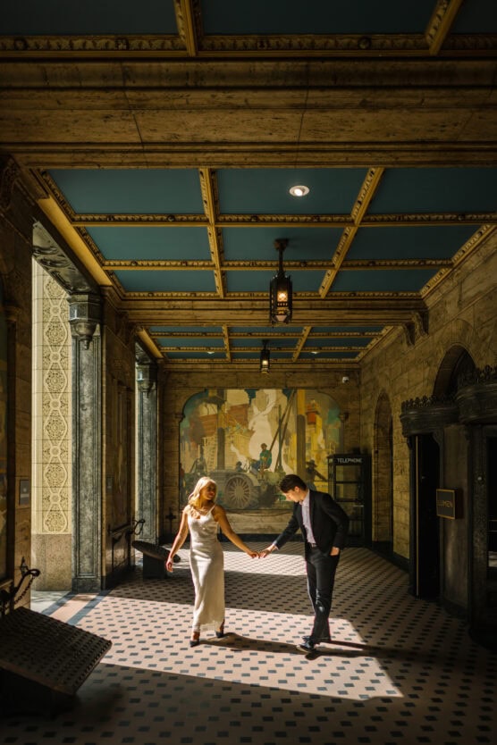 A couple stands in an ornate hallway with checkered flooring and a painted mural. The woman in a white dress holds hands with the man in a dark suit as sunlight streams in, creating a warm, elegant atmosphere. by Justin Salem Meyer