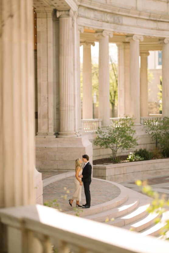 A couple stands closely together on stone steps in front of a grand, columned building, surrounded by greenery and soft sunlight. by Justin Salem Meyer