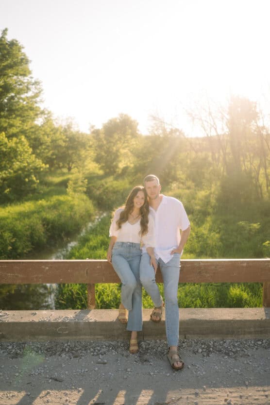 A couple sits together on a wooden railing in the sunlight, surrounded by green trees and grass. They both wear light-colored shirts and jeans, smiling at the camera on a bright, sunny day. by Justin Salem Meyer