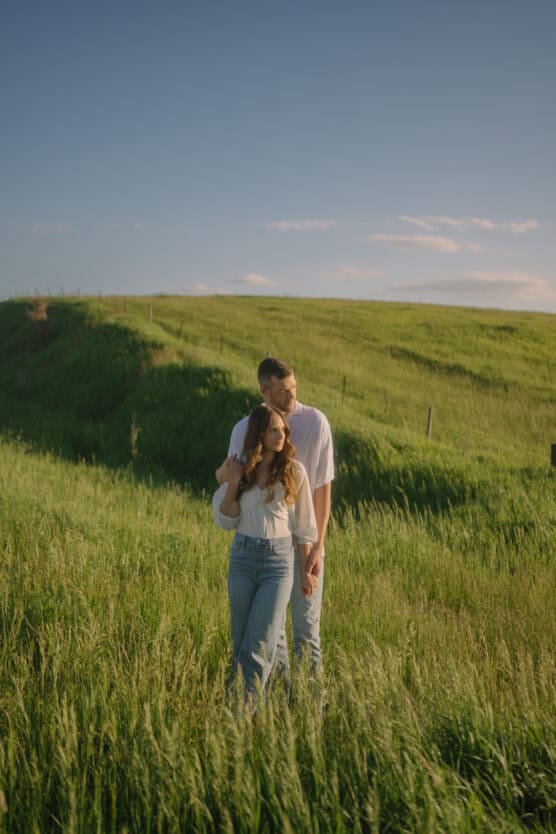 A couple stands closely together in a sunlit green field, with rolling grassy hills and a blue sky with light clouds in the background. The woman looks ahead while the man gently holds her from behind. by Justin Salem Meyer
