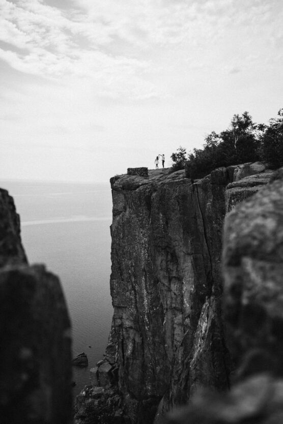 Two people stand together near the edge of a tall, rocky cliff overlooking a calm body of water, with trees nearby and a dramatic sky above in black and white. by Justin Salem Meyer