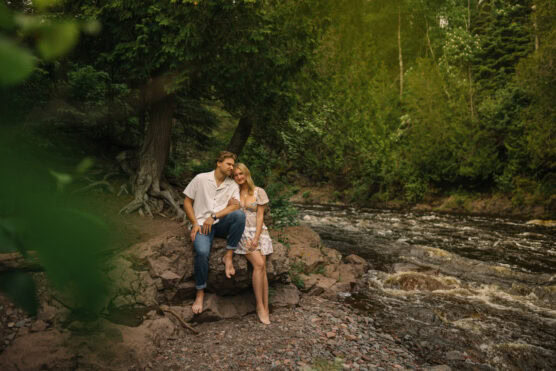 A couple stands barefoot on rocks by a flowing river, surrounded by lush green trees. The man leans against a tree while the woman stands close, both looking content and relaxed in the natural woodland setting. by Justin Salem Meyer
