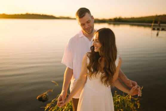 A couple stands by a lake at sunset, smiling and holding hands. The woman wears a white dress and has long brown hair; the man stands behind her in a white shirt. The scene is warm and romantic with golden sunlight. by Justin Salem Meyer