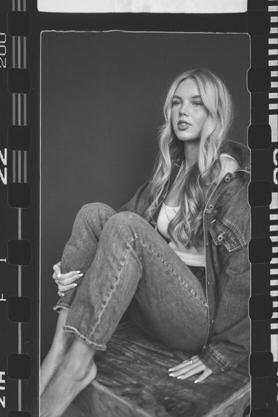 A black and white photo of a woman with long, wavy hair, wearing a denim jacket and jeans, sitting barefoot on a wooden box against a plain dark background. The image is framed with film strip borders. by Justin Salem Meyer