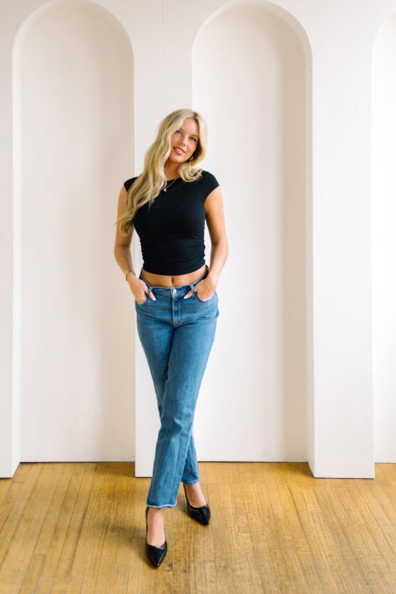 A woman with long blonde hair stands indoors against a white wall with arched cutouts. She wears a black fitted t-shirt, blue jeans, and black heels, with her hands in her pockets and a relaxed, smiling expression. by Justin Salem Meyer