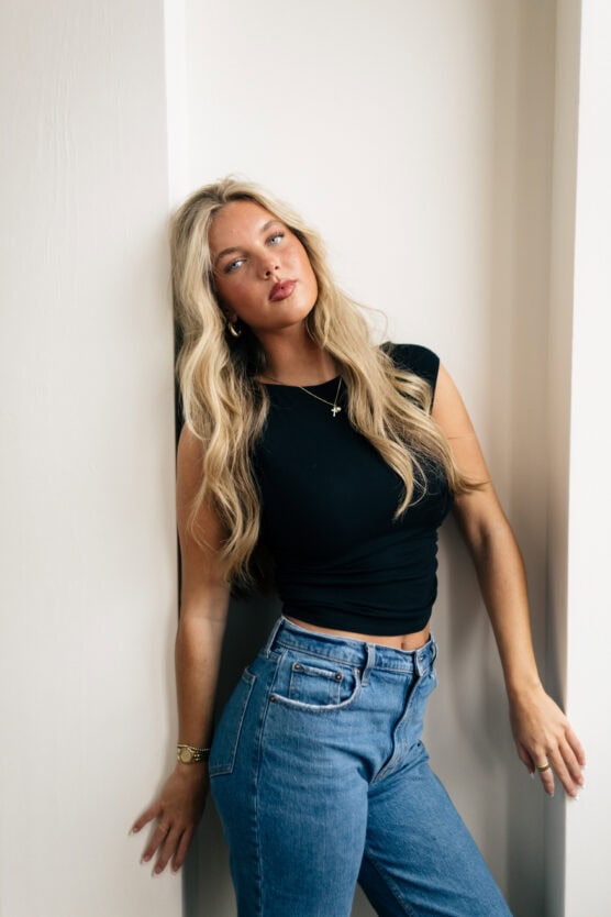 A woman with long blonde hair wearing a black sleeveless top and blue jeans leans against a white wall, looking confidently at the camera with a relaxed posture. by Justin Salem Meyer