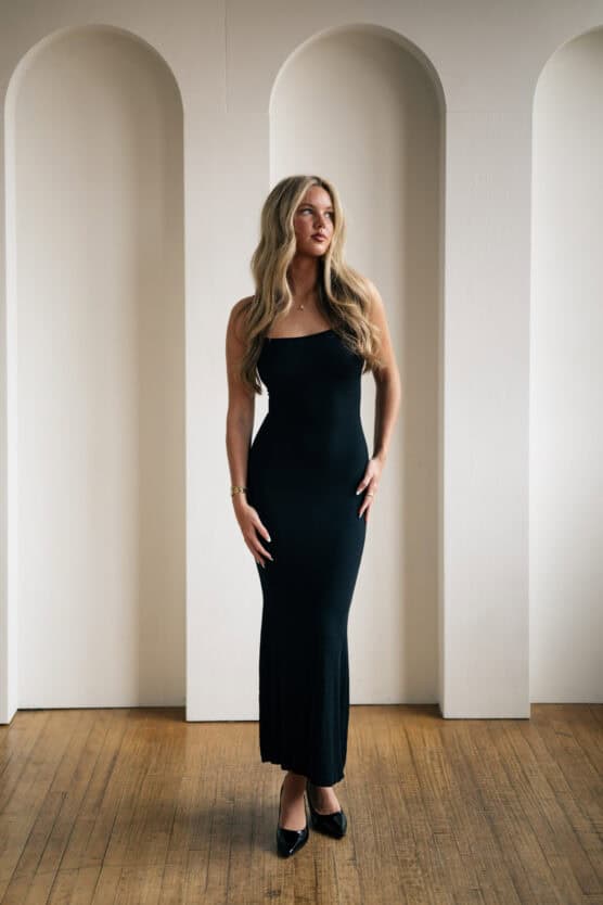A woman with long blond hair wearing a sleeveless black dress and black heels stands on a wooden floor in front of white arched wall panels, looking slightly to the side. by Justin Salem Meyer
