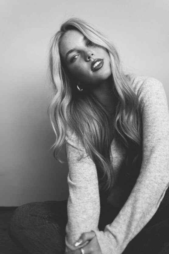 A woman with long, wavy hair poses seated against a plain background, looking slightly up at the camera. She wears a light sweater and hoop earrings. The photo is in black and white. by Justin Salem Meyer