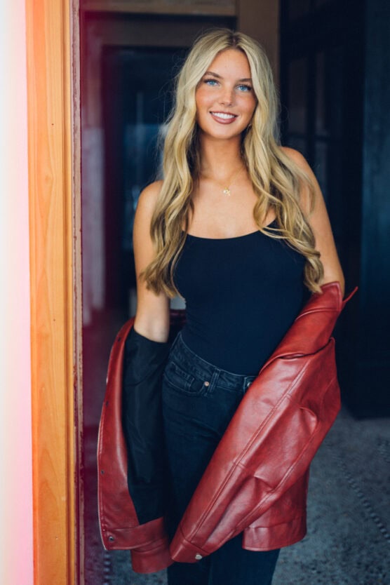A young woman with long blonde hair, wearing a black tank top and dark jeans, stands indoors holding a red leather jacket open with both hands, smiling at the camera. by Justin Salem Meyer