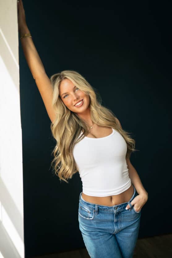 A smiling woman with long blonde hair wearing a white tank top and blue jeans stands against a dark background, with one arm raised and resting on a white wall. by Justin Salem Meyer