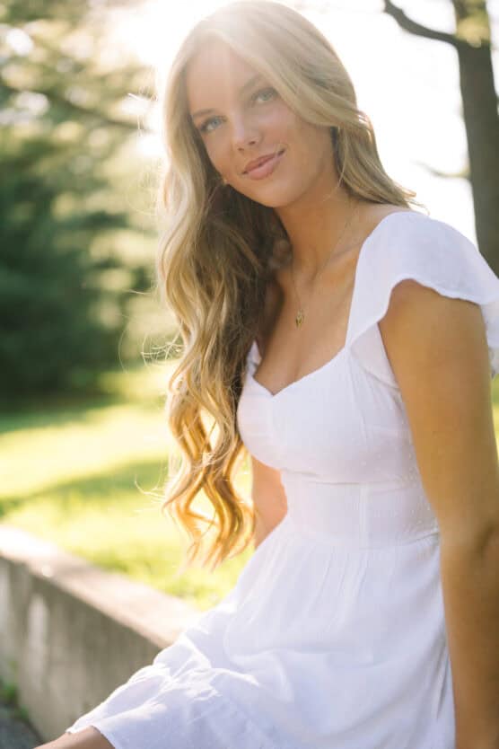 A young woman with long, wavy blonde hair sits outdoors in sunlight, wearing a white dress with short sleeves. She smiles softly, with a blurred background of green trees and grass. by Justin Salem Meyer