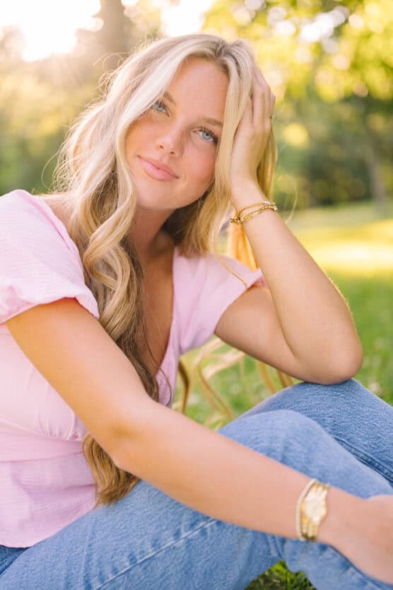 A young woman with long blonde hair sits on grass in sunlight, wearing a pink top and blue jeans, smiling softly with her hand resting on her head in a relaxed, outdoor setting. by Justin Salem Meyer
