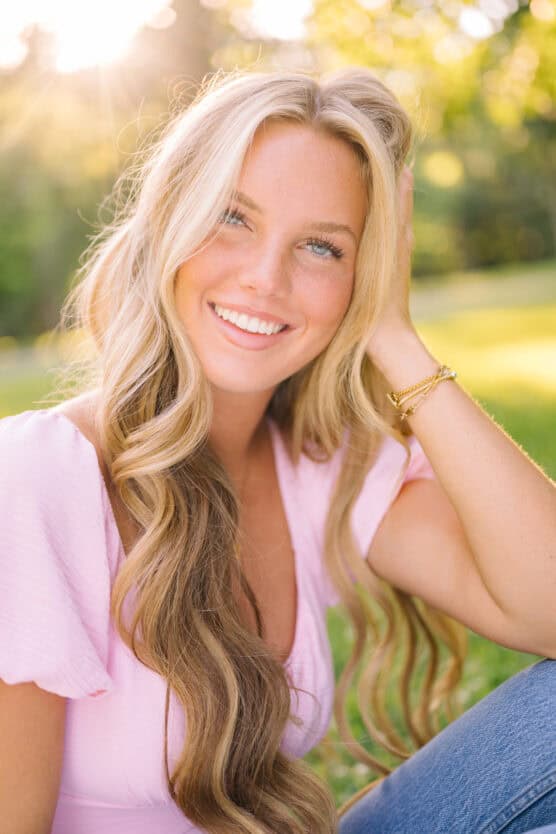 A young woman with long, wavy blonde hair sits outdoors on grass, smiling at the camera. She wears a light pink top and blue jeans, with sunlight softly filtering through green trees behind her. by Justin Salem Meyer