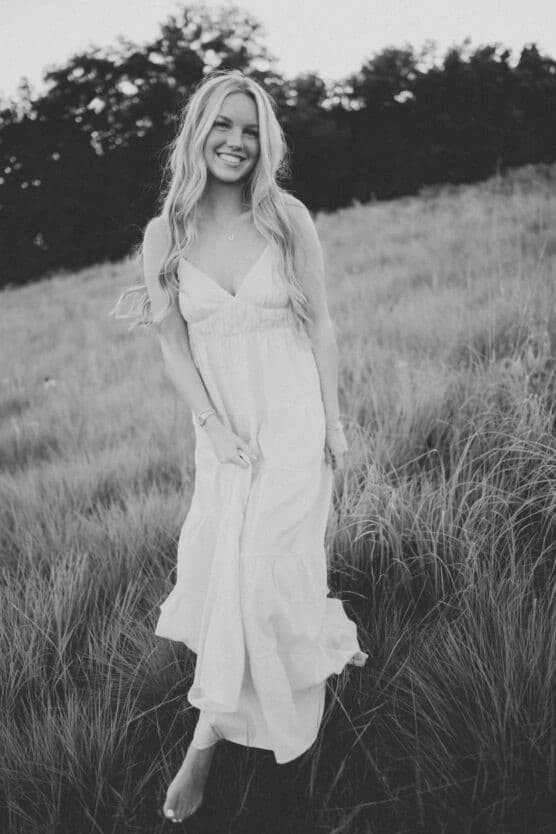 A woman in a long, flowing dress stands barefoot in a grassy field, smiling at the camera. Trees are visible in the background. The image is in black and white. by Justin Salem Meyer
