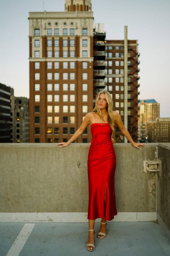 A woman in a strapless red dress and heels stands on a rooftop parking lot, with tall city buildings in the background during sunset. by Justin Salem Meyer
