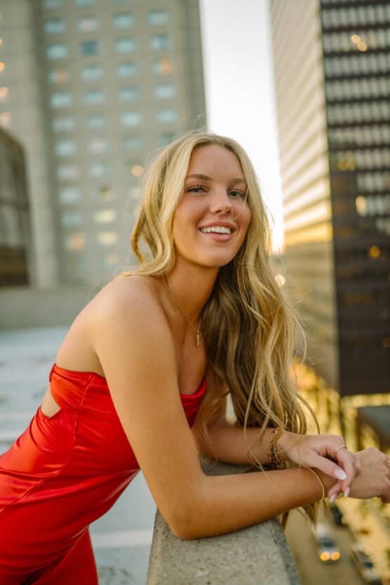 A young woman with long blonde hair in a red dress leans on a concrete ledge, smiling, with tall buildings and city lights in the blurred background at sunset. by Justin Salem Meyer