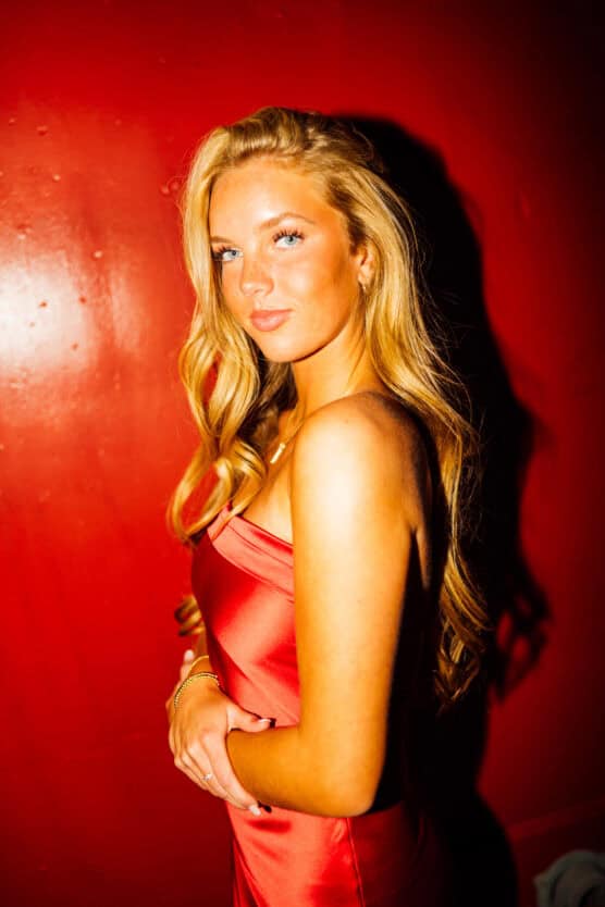 A woman with long blonde hair wearing a red satin dress stands against a bright red wall, looking at the camera with a confident expression. Warm lighting highlights her features and the texture of her dress. by Justin Salem Meyer