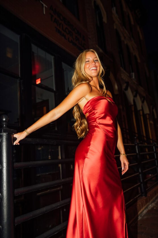 A woman in a shiny red strapless gown stands outside at night, smiling and leaning on a black metal railing, with buildings and a lit doorway in the background. by Justin Salem Meyer