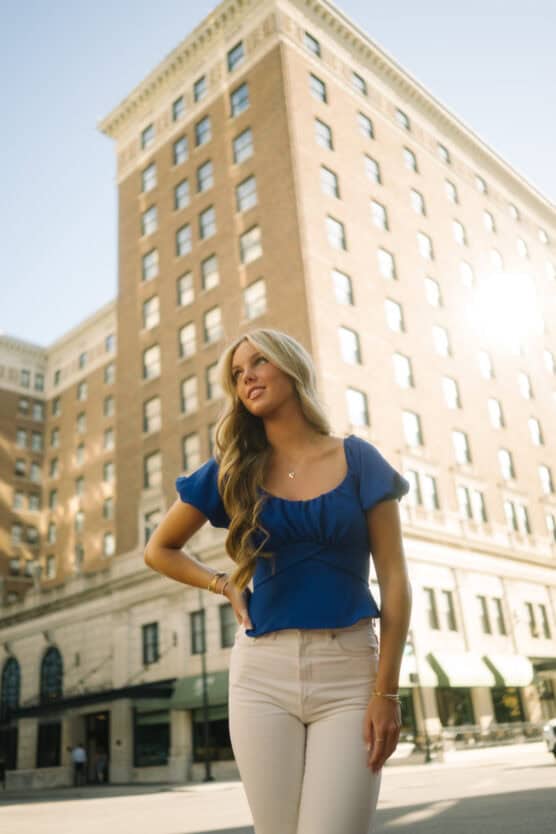 A young woman with long blonde hair, wearing a blue top and white pants, stands outdoors in front of a tall brick building on a sunny day. by Justin Salem Meyer