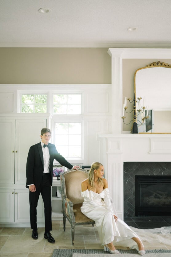 A man in a black tuxedo stands beside a woman in a white off-the-shoulder dress seated on a chair in an elegant room with white cabinets, a fireplace, and a large mirror above the mantel. by Justin Salem Meyer