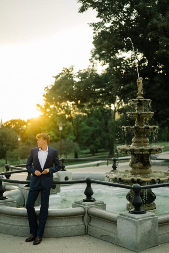 A person in a blue suit stands by an ornate stone fountain with water spraying, surrounded by trees at sunset, with sunlight shining in the background. by Justin Salem Meyer