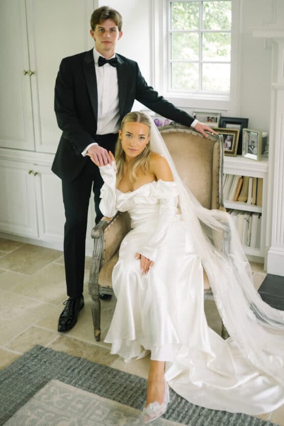 A bride in a long white off-the-shoulder gown and veil sits on a chair, holding hands with a groom standing beside her in a black tuxedo and bow tie. They pose in a bright room with white walls and bookshelves. by Justin Salem Meyer