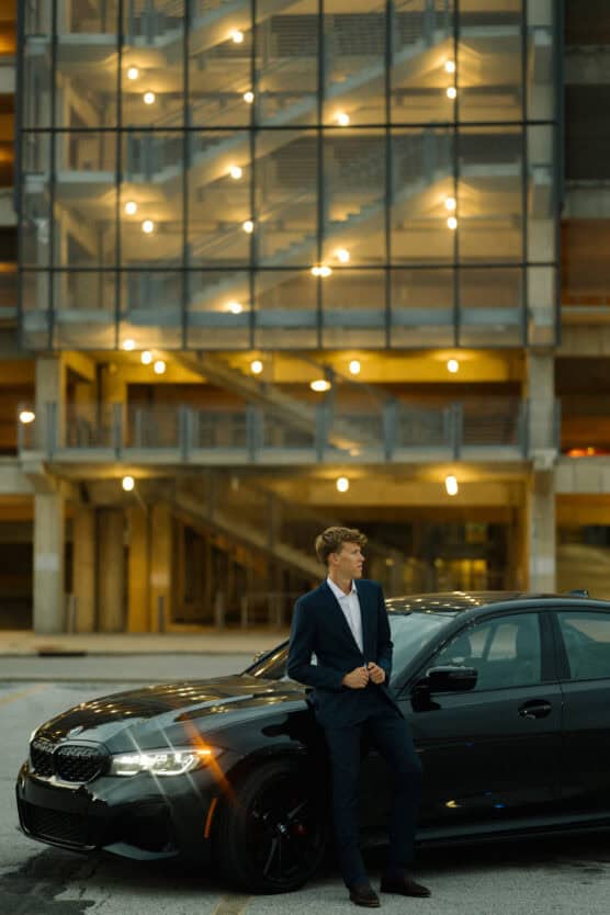 A man in a dark suit stands leaning against a black car in front of a modern building with illuminated stairways and glass panels at dusk. by Justin Salem Meyer