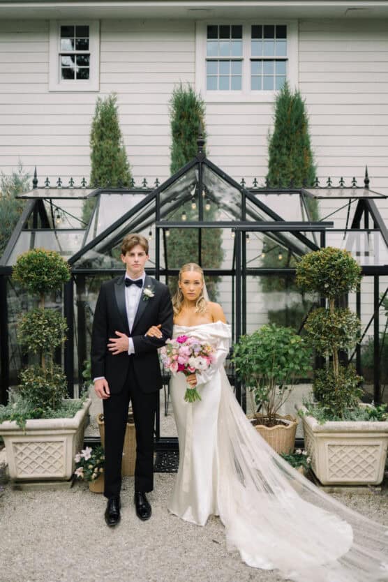 A bride in a white gown and veil holding a bouquet stands beside a groom in a black tuxedo in front of a glass greenhouse, with potted plants and a white building in the background. by Justin Salem Meyer
