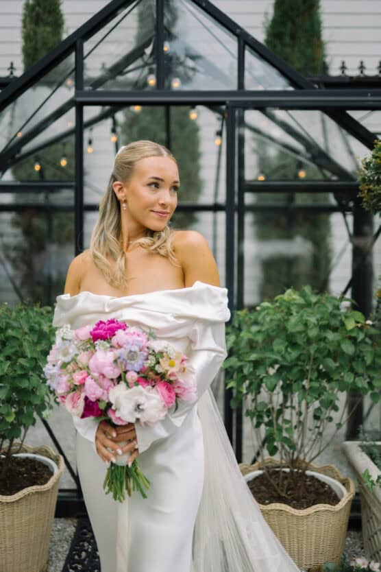 A bride in an off-the-shoulder white dress holds a colorful bouquet of pink and white flowers, standing in front of a glass greenhouse with lush greenery and string lights. by Justin Salem Meyer